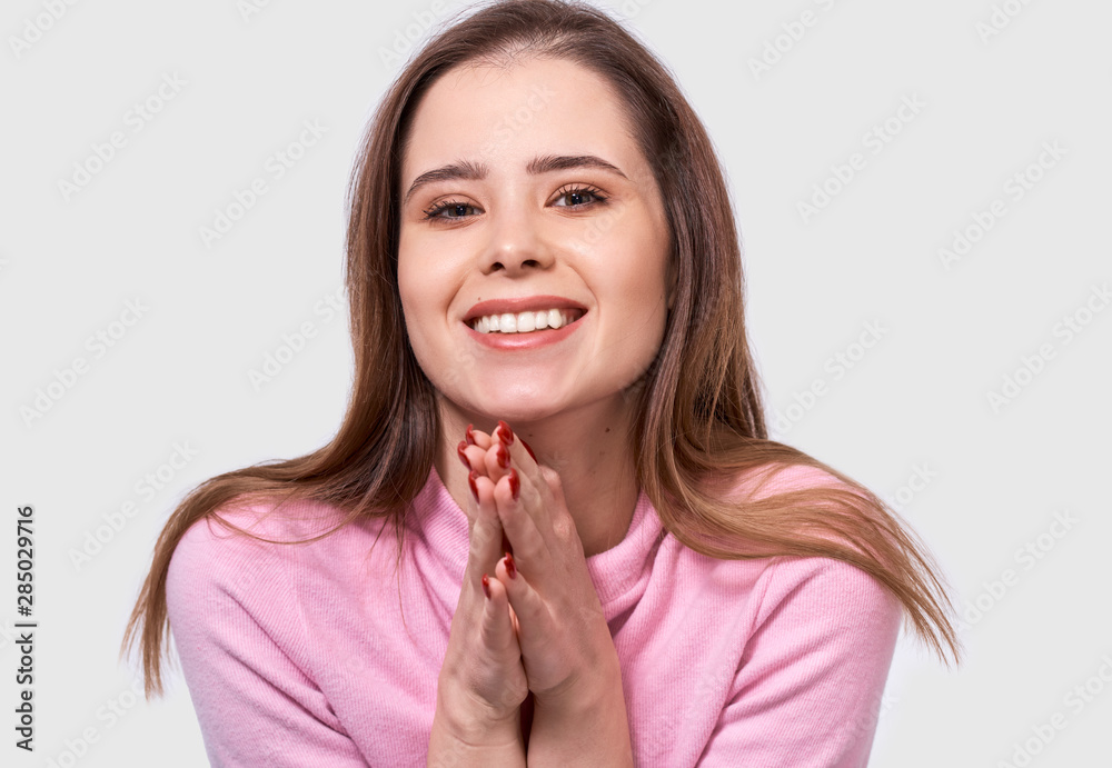 Closeup portrait of happy young woman smiling broadly, holding both hands on the chin, looking to the camera, has joyful expression. Charming Caucasian female has positive emotions. Closeup portrait of happy young woman smiling broadly, holding both hands on the chin, looking to the camera, has joyful expression. Charming Caucasian female has positive emotions.