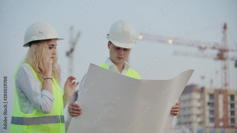 Two builders with drawings standing on the background of buildings under construction in helmets and vests, a woman talking on the phone with the customer Two builders with drawings standing on the background of buildings under construction in helmets and vests, a woman talking on the phone with the customer