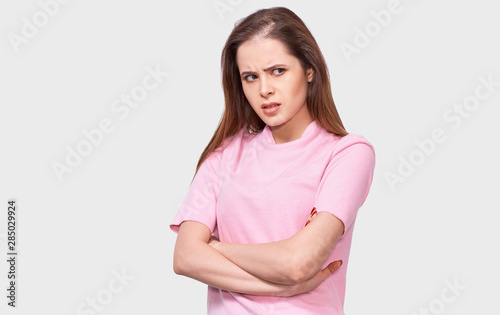 Mad young woman has upset facial expression, wears pink casual outfit, keeps crossed hands on waist, isolated over white studio background. Upset female looking aside, posing over gray studio wall. © iuricazac