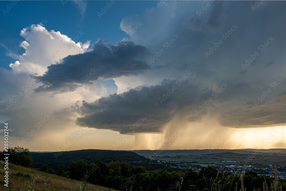 Dunkle Gewitterwolken und aufstseigende weiße Wolken mit letztem blauen Himmel und Regen bei Sonnenschein am Horizont über Weißenburg in Bayern