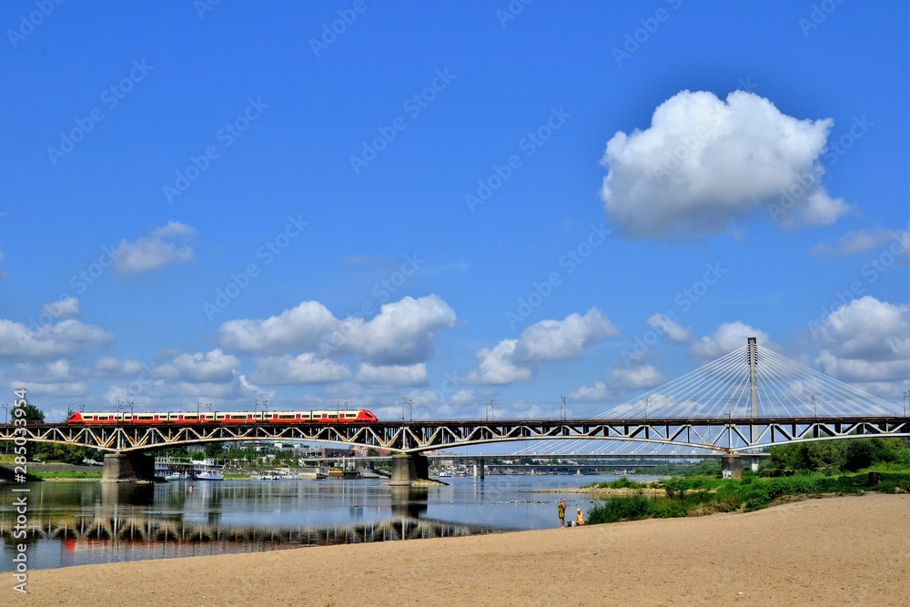 The red train crossing the Srednicowy Bridge in Warsaw, Poland. View from the Stadium Beach (or Poniatowka beach) in the vicinity of the National Stadium. 