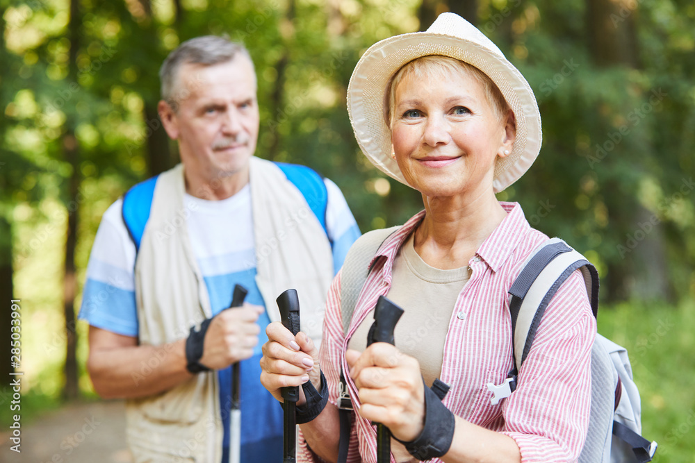 Portrait of mature woman in hat and with backpack looking at camera while doing nordic walking together with her husband in the nature