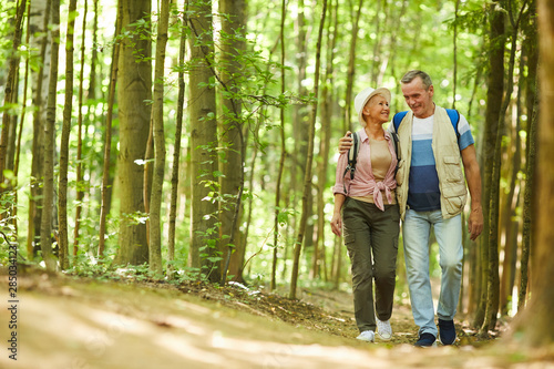 Senior happy couple embracing each other and walking along the footpath during their hike in the forest in summer day