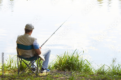 Rear view of mature fisherman sitting on chair with fishing rod and waiting for a catch at the lakeside outdoors
