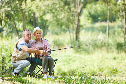 Mature man helping to hold the woman fishing rod while she sitting on chair and fishing near the pond outdoors