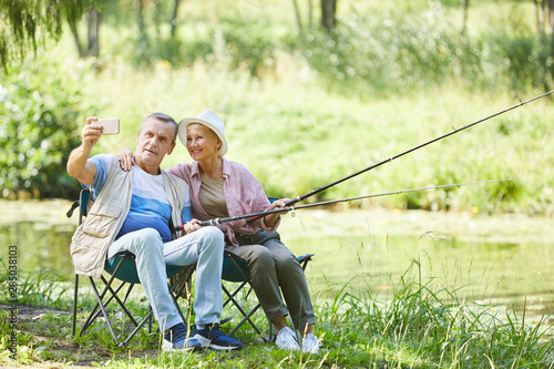 Happy mature couple making a selfie portrait on mobile phone while fishing together near the river in nature