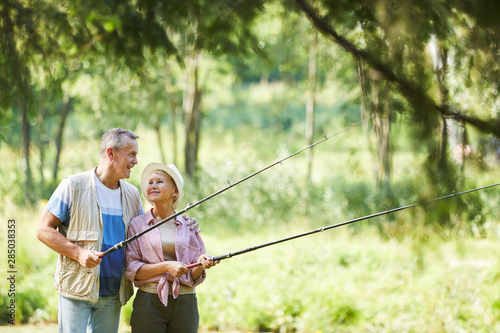 Mature couple standing and talking to each other while holding fishing rods and fishing together outdoors