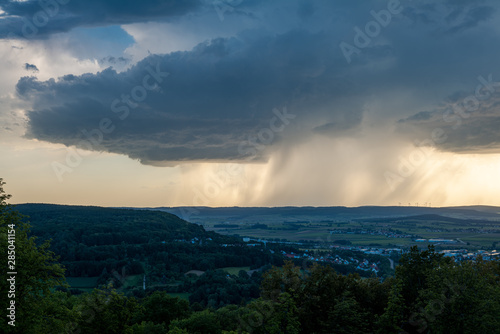 Dunkle Gewitterwolken mit letztem blauen Himmel und Regen bei Sonnenschein am Horizont über Weißenburg in Bayern
