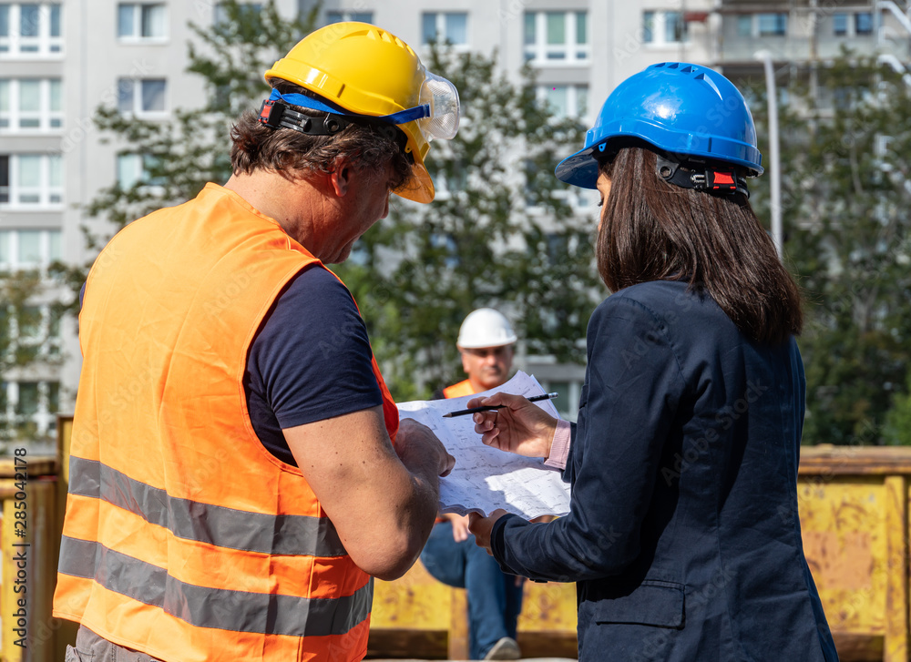 Construction manager wearing safety jacket and helmet checking projects discussing with a female engineer. In background, another worker Construction manager wearing safety jacket and helmet checking projects discussing with a female engineer. In background, another worker