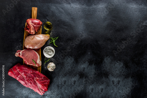 Various meat types. Red beef meat, pork steak, chicken breast filet, liver pork heart, with spices on black stone concrete background top view copy space