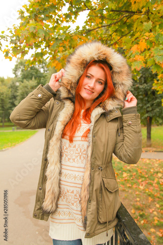 Redheaded woman in coat with fur hood in autumn park with yellow green trees