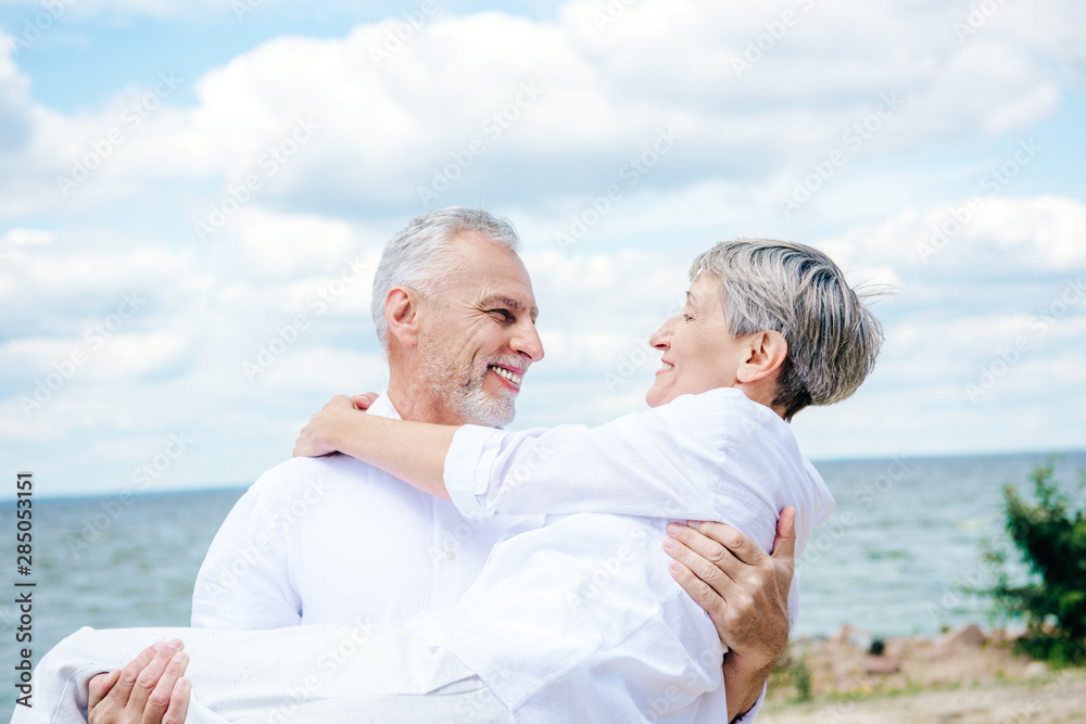smiling senior man in white shirt lifting wife under blue sky