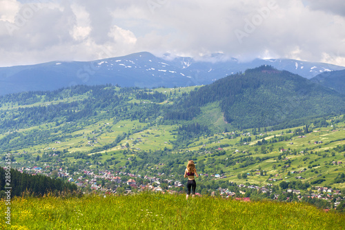 Back view of slim young woman standing on grassy valley on background of green mountains on sunny summer day. © bilanol