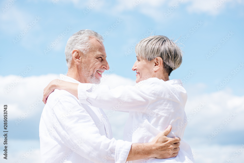 side view of smiling senior couple embracing and looking at each other under blue sky side view of smiling senior couple embracing and looking at each other under blue sky