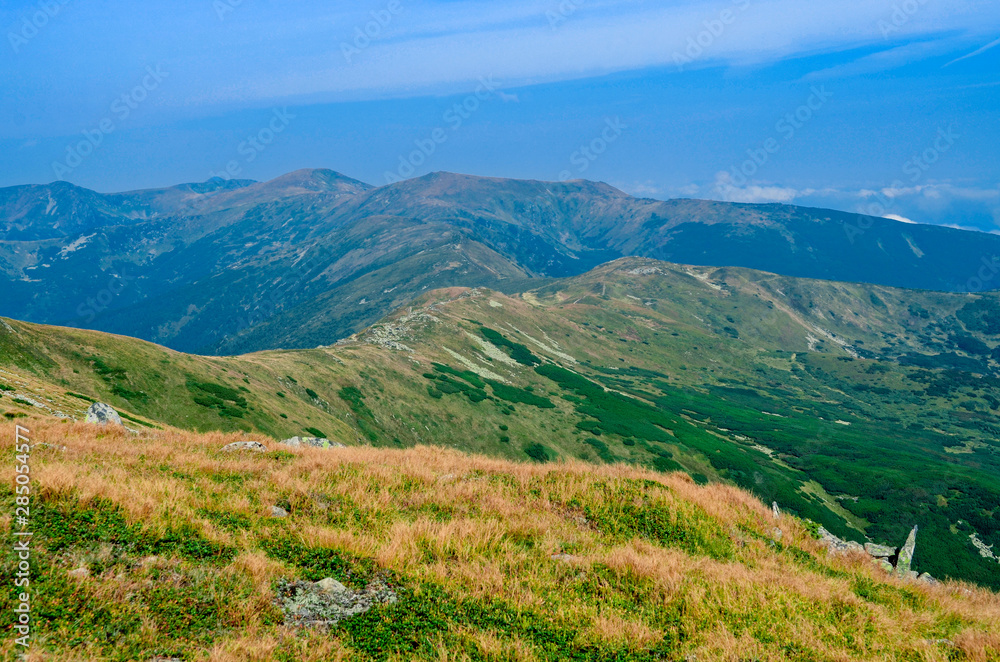The high mountain range is overgrown with green and yellow grass against the blue sky with clouds