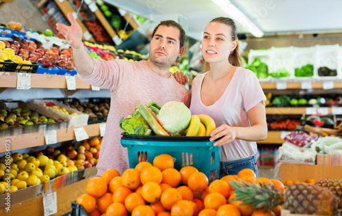 Positive family couple standing with full cart after shopping and pointing to shelves in fruit store