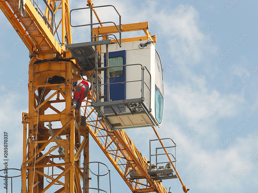 Crane workers in overalls are repairing a high-rise tower crane working during the construction of a multi-storey building. Risky work at height. Lifting heavy building materials. City development. Crane workers in overalls are repairing a high-rise tower crane working during the construction of a multi-storey building. Risky work at height. Lifting heavy building materials. City development.
