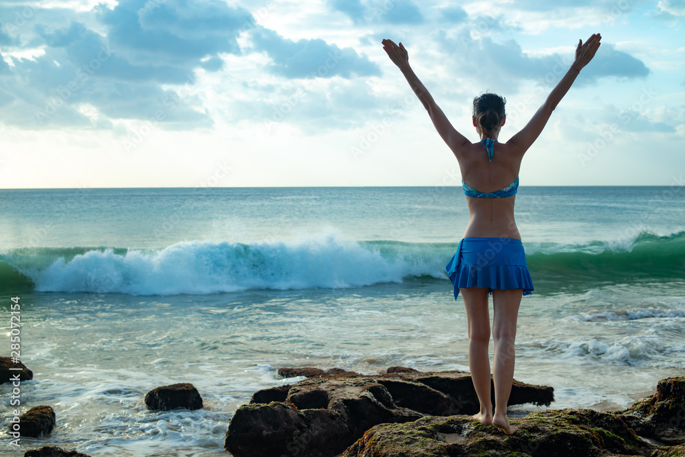 Excited young woman standing on the rock, raising arms at the beach in front of the ocean. View from back. Sunset at the beach. Luxury travel to Asia. Ocean waves. Copy space. Tegal Wangi, Bali