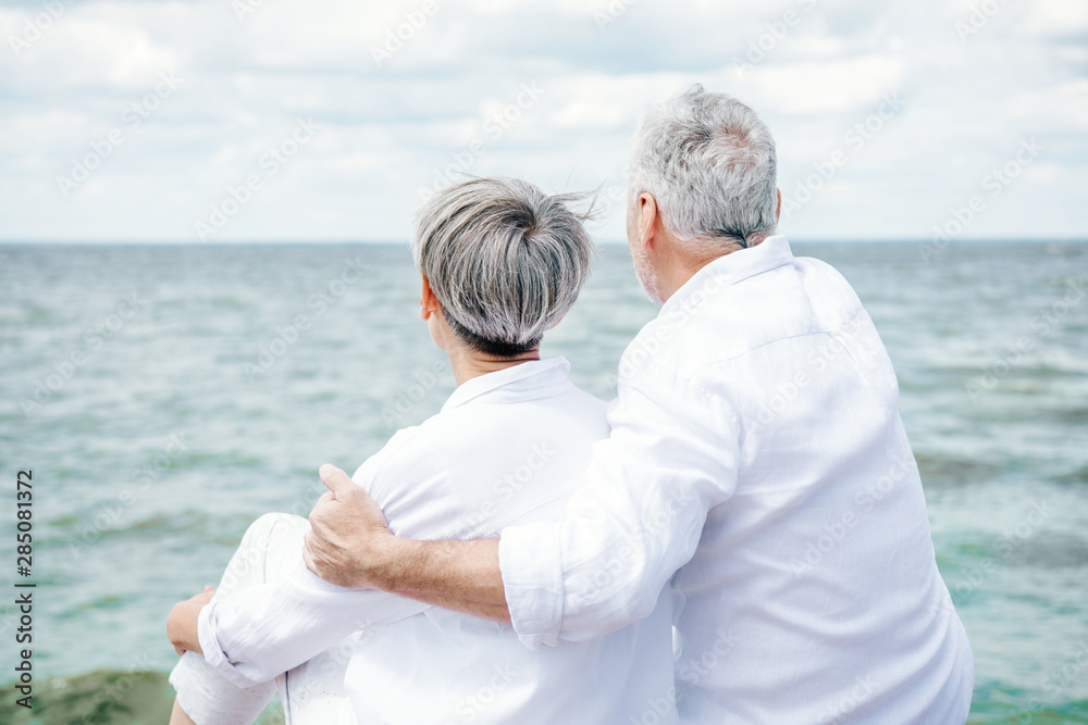 back view of senior couple in white shirts embracing near river under blue sky