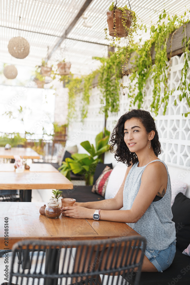 Vertical shot modern stylish young brunette woman with curly short hair sitting alone cafe lounge area, summer terrace, contemplate tropical island hotel scenery, smiling sensual, drink cappuccino Vertical shot modern stylish young brunette woman with curly short hair sitting alone cafe lounge area, summer terrace, contemplate tropical island hotel scenery, smiling sensual, drink cappuccino