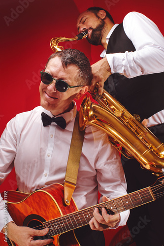 Group of two musicians, male jazz band, guitarist and saxophonist in classical costumes improvise on musical instruments in a studio on  red background