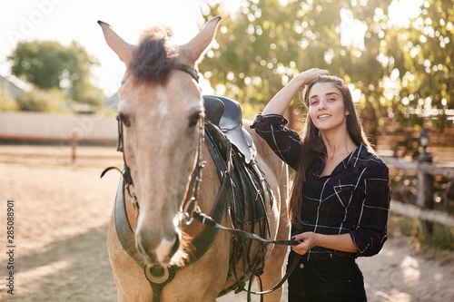 Charming equestrian, female rider standing with her favorite brown cute horse, holding bridle, put saddle, wanna take ride in morning, spend time on family rancho, smiling joyfully