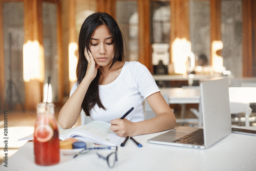 Concentration, digital nomad and internet education concept. Pensive beautiful young woman sit cafe drink juice and writing down important information from university web-page using laptop Concentration, digital nomad and internet education concept. Pensive beautiful young woman sit cafe drink juice and writing down important information from university web-page using laptop