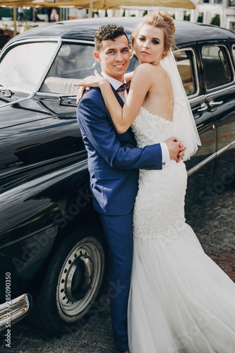 Stylish happy bride and groom embracing at old black retro car. Gorgeous wedding couple of newlyweds hugging after wedding ceremony. Romantic moment © sonyachny