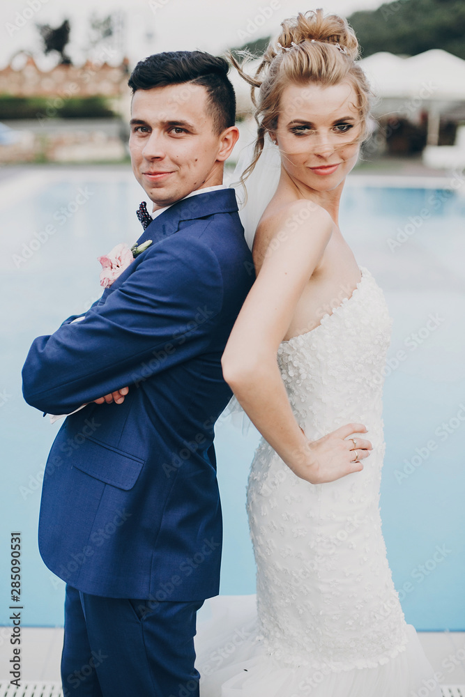 Naklejka premium Stylish happy bride and groom posing at pool blue water at wedding reception in restaurant. Gorgeous wedding couple of newlyweds having fun after wedding ceremony outdoors