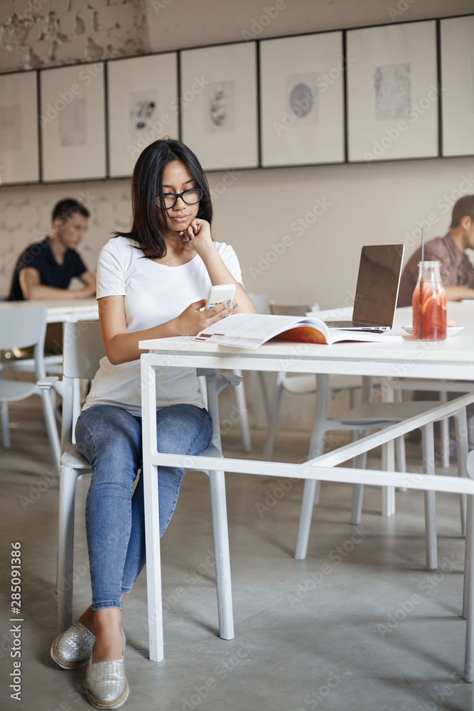 Vertical shot busy young woman in glasses sit alone cafe surrounded with working material notebook and laptop, using smartphone, scrolling social media feed during break from studying Vertical shot busy young woman in glasses sit alone cafe surrounded with working material notebook and laptop, using smartphone, scrolling social media feed during break from studying