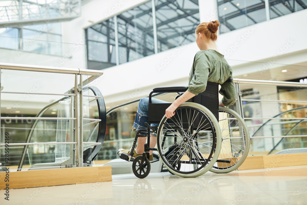 Rear view of disabled young woman going in wheelchair she standing near the escalator in shopping centre Rear view of disabled young woman going in wheelchair she standing near the escalator in shopping centre