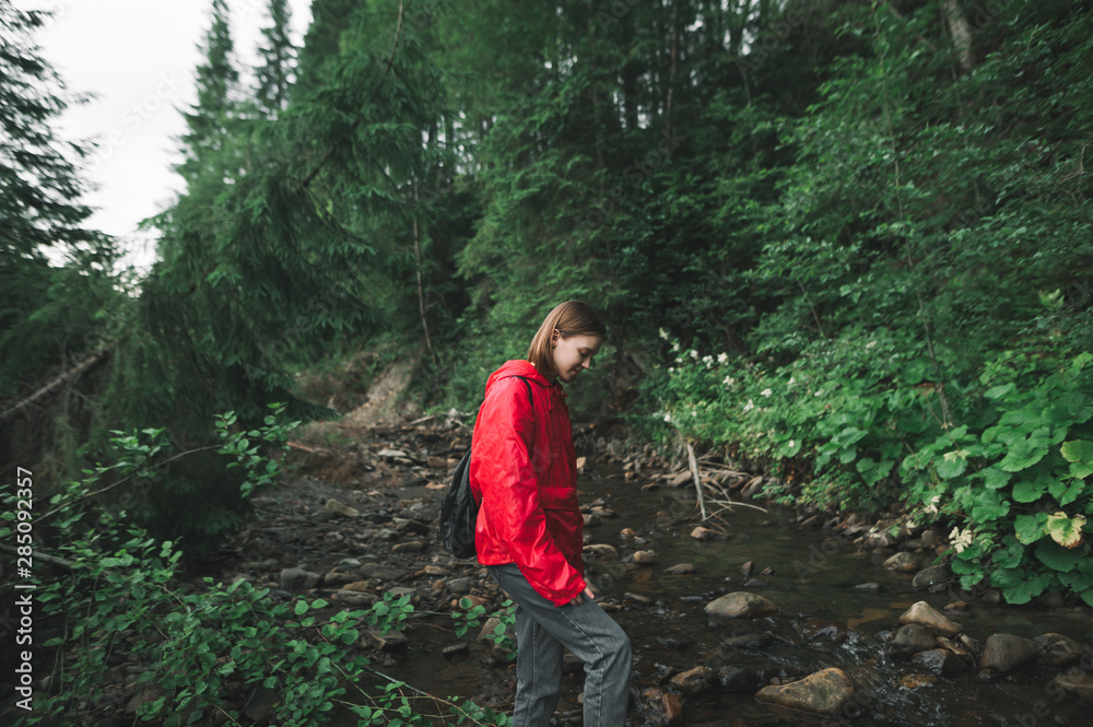 Beautiful hiker girl in a red raincoat walks in the forest in the wild,crosses a mountain stream, looks down at the water. Hiking woman in the mountains, vacation in the wild.