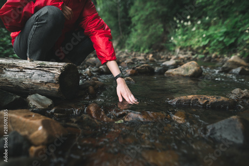 Hiker woman lowers her hand into a clear cold, mountain stream, close up photo. Hiker girl washes her hands in a mountain river. Background