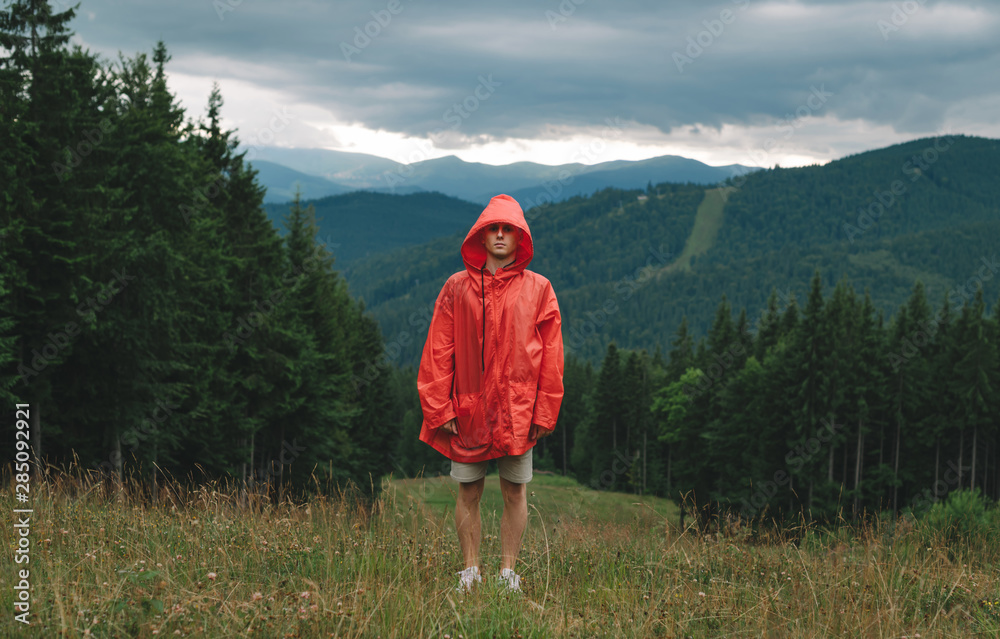 Full length portrait, young man in red raincoat stands in mountains in bad rainy weather, on coniferous forest background, looks into camera with serious face.Guy tourist on a mountain hike poses