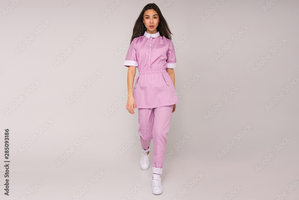 young stylish nurse in pink medical costume with long brunette hair is standing, looking straight and smiling cute on white wall background. medical fashion concept. free space young stylish nurse in pink medical costume with long brunette hair is standing, looking straight and smiling cute on white wall background. medical fashion concept. free space