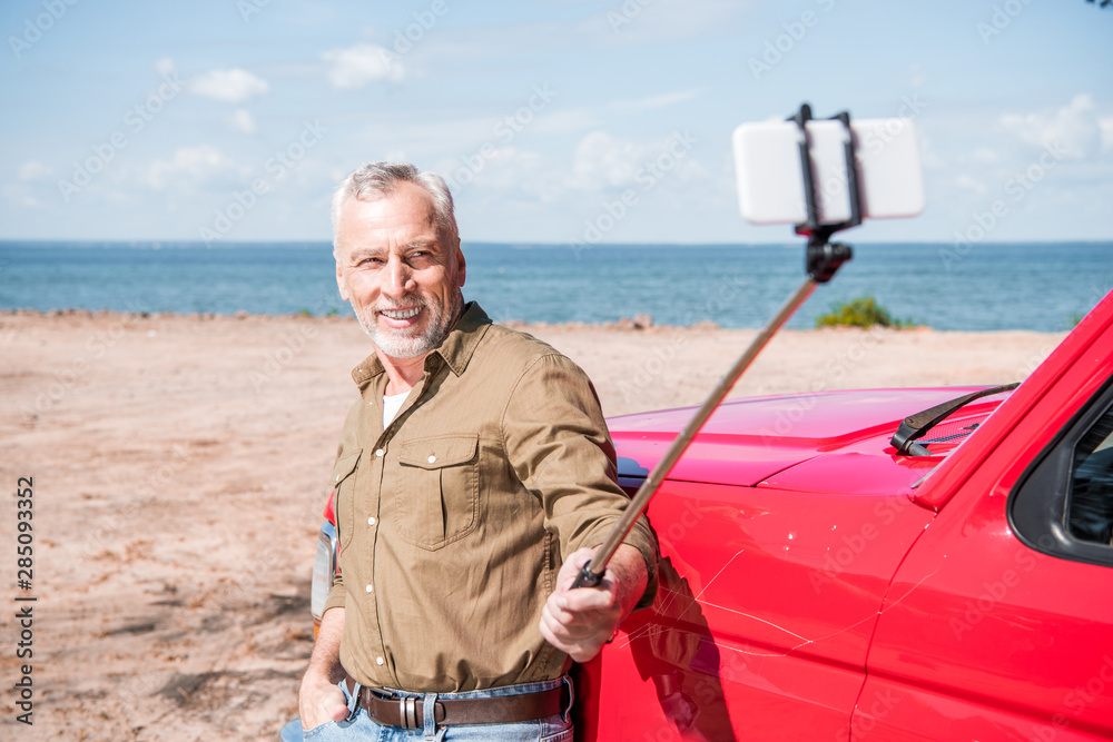 Fototapeta premium smiling senior man standing near red car and taking selfie in sunny day