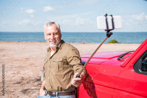 smiling senior man standing near red car and taking selfie in sunny day
