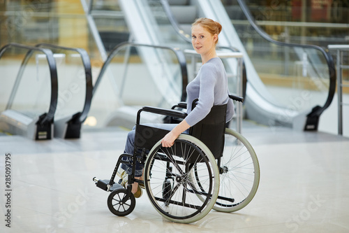 Portrait of young red-haired woman sitting in wheelchair and looking at camera while going along the shopping mall © AnnaStills