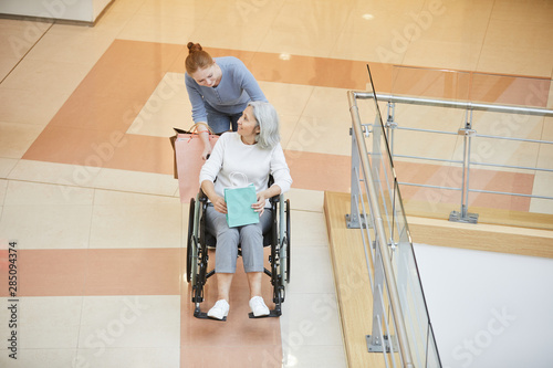 Young daughter talking to her senior disabled mother in wheelchair while they going along the corridor in shopping mall