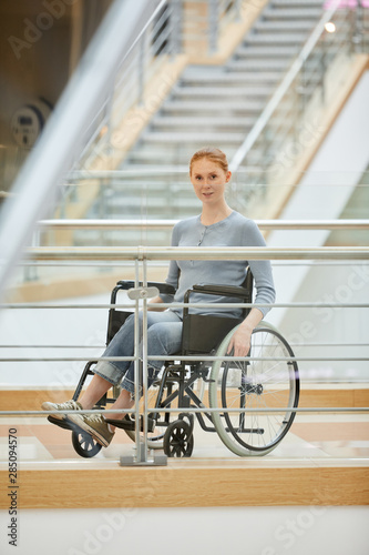Portrait of young disabled woman in casual cloting sitting in wheelchair and looking at camera at modern hospital