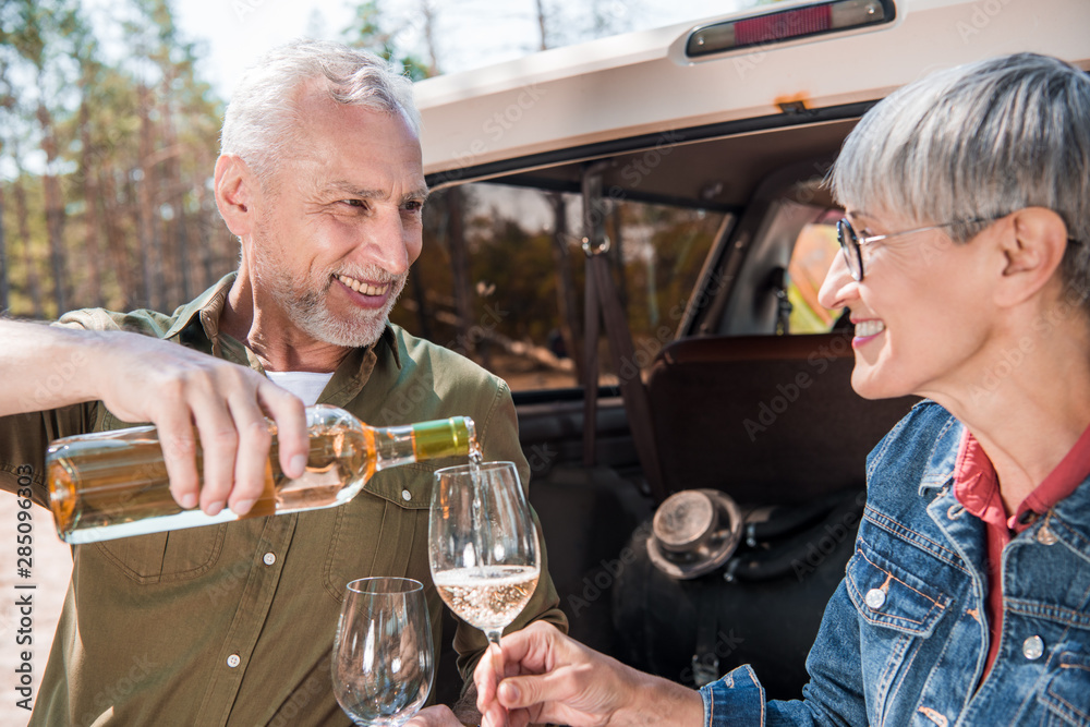 smiling senior man pouring wine in wine glasses and looking at wife near car