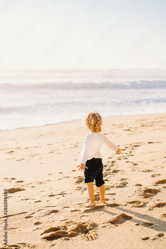 Barefoot little boy with blond curly hair standing on the beach and watching the ocean view in California. Traveling with kids.