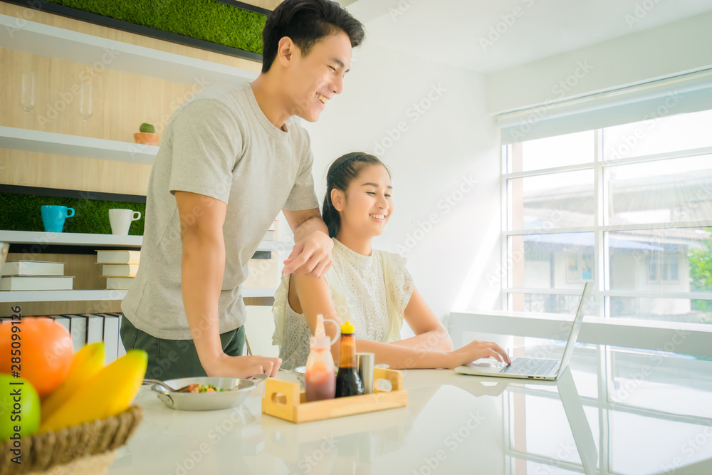 Asian Handsome man 24 years old smile and stand beside with Asian beautiful woman 24 years old smile, sitting both used laptop search for cooking methods on table in home in the morning near window