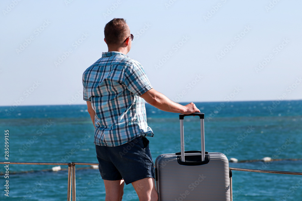 Fototapeta premium Tourist and traveler is stand on beach of hotel resort with traveled suitcase, enjoys view sea environment, nature landscape, panorama, horizon and rest in sun. Handsome man on vacation in summertime.