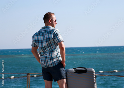 Tourist and traveler is stand on beach of hotel resort with traveled suitcase, enjoys view sea environment, nature landscape, panorama, horizon and rest in sun. Handsome man on vacation in summertime.