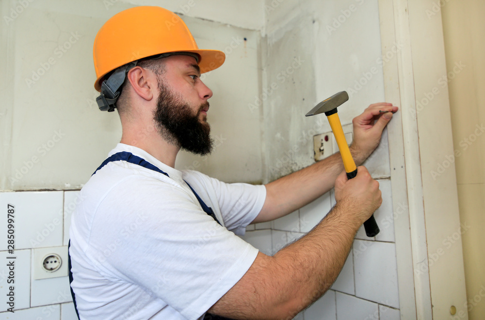 Construction worker and handyman working on renovation work. Builder with  yellow hammer strikes and nails a nail into wooden wall of kitchen door at  construction site. Apartment renovation concept. Stock Photo |
