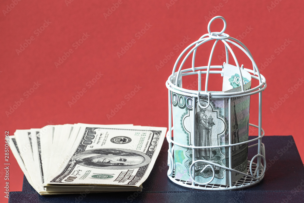 the ruble closed in a tight cage and a pack of dollars lying next to the book, the background is red the ruble closed in a tight cage and a pack of dollars lying next to the book, the background is red