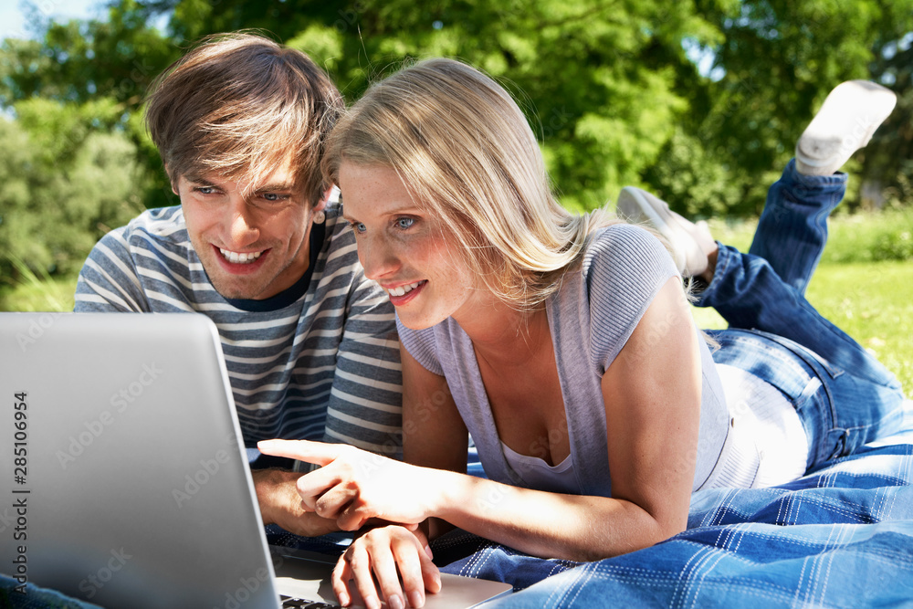 Germany, Cologne, Young couple using laptop in meadow, smiling Germany, Cologne, Young couple using laptop in meadow, smiling