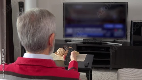 An elderly man watches out-of-focus TV and switch programs with a remote control in a living room - view from behind