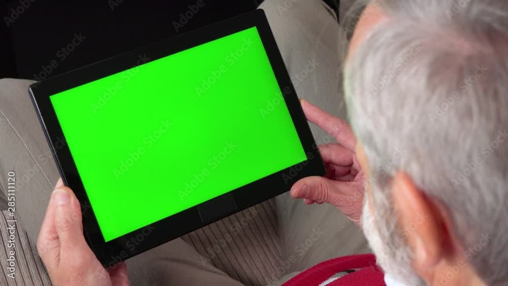 An elderly man sits on a couch in an apartment and looks at a tablet with a green screen - closeup from behind his head